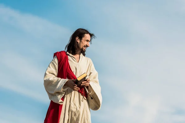 Jesus holding cross and holy bible against sky with clouds — Stock Photo