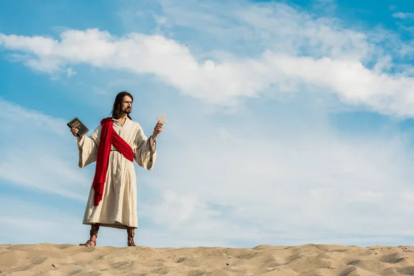 Jesus mit heiliger Bibel und Kreuz vor blauem Himmel mit Wolken in der Wüste — Stockfoto