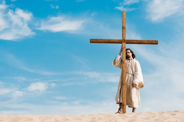 Bearded man standing near wooden cross in desert on golden sand — Stock Photo