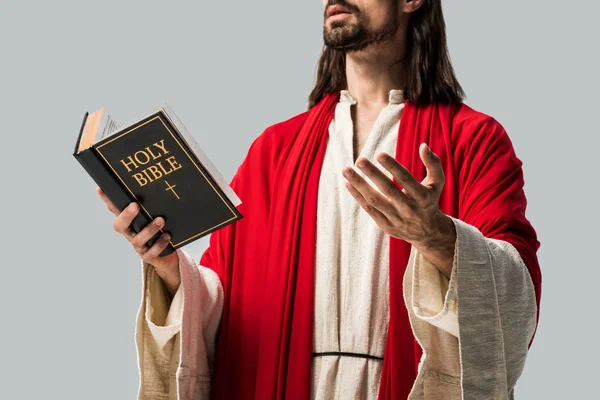 Vista ritagliata di uomo religioso in vestaglia Gesù tenendo santa bibbia e gesticolando isolato su grigio — Foto stock