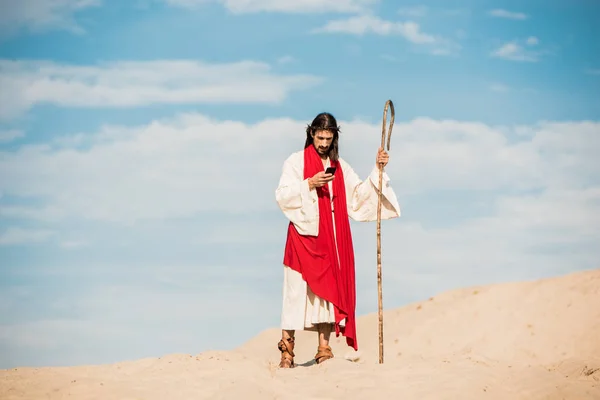 Handsome man in jesus robe holding wooden cane and using smartphone in desert — Stock Photo