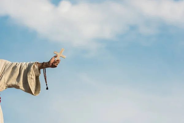 Cropped view of religious man holding cross and rosary beads against blue sky — Stock Photo