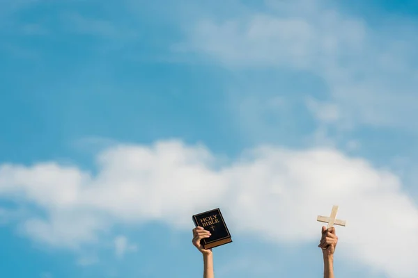 Обрізаний вид людини, що тримає святу Біблію і хрест на блакитне небо з хмарами — стокове фото