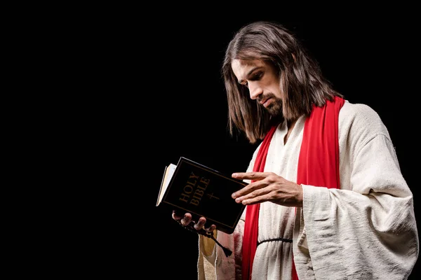 Hombre religioso sosteniendo libro con las letras sagradas de la biblia aisladas en negro - foto de stock