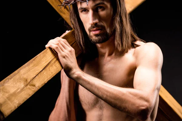 Religious and shirtless man in wreath with spikes holding wooden cross isolated on black — Stock Photo