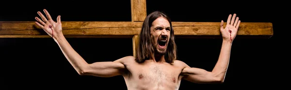 Panoramic shot of shirtless man crucified on wooden cross screaming isolated on black — Stock Photo