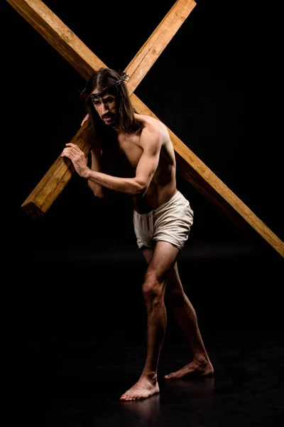 Иисус в венке, держа тяжелый деревянный крест и ходить по черному — стоковое фото