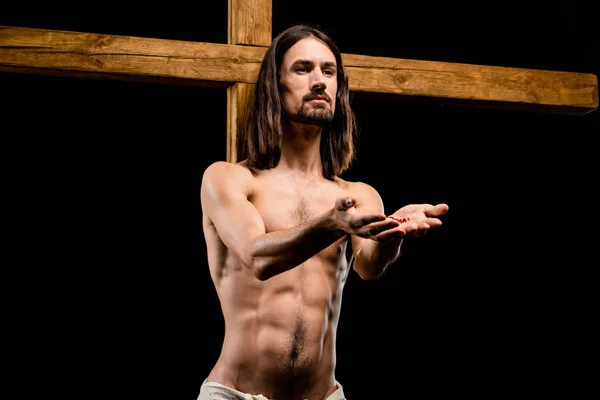 Jesus with cupped hands near wooden cross isolated on black — Stock Photo