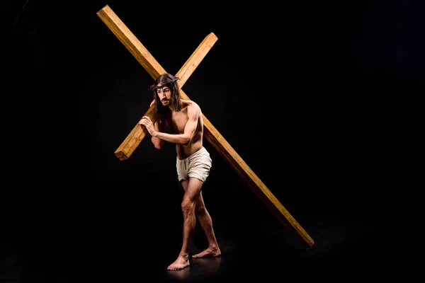 Torse nu Jésus en couronne tenant une croix en bois et marchant sur le noir — Photo de stock
