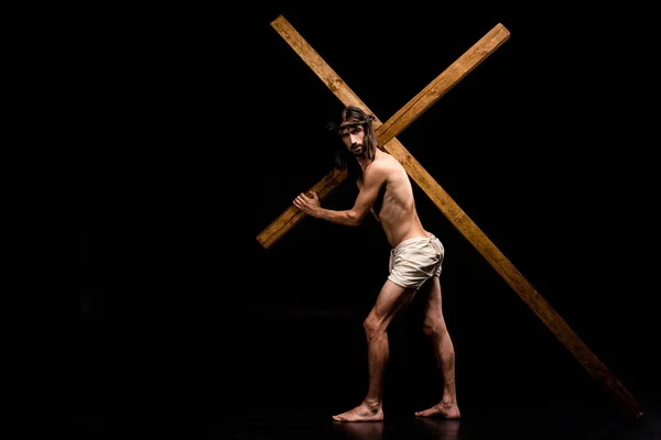 Shirtless jesus in wreath holding wooden cross on black — Stock Photo
