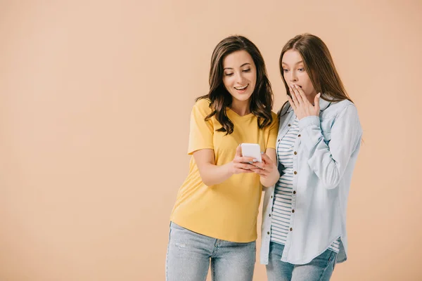 Mujer atractiva mirando el teléfono inteligente con su amigo sorprendido aislado en beige - foto de stock