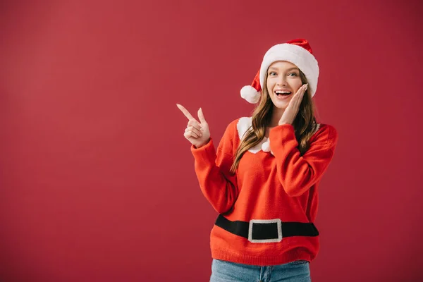 Attractive woman in santa hat and sweater pointing with finger isolated on red — Stock Photo