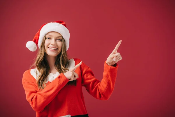 Attractive woman in santa hat and sweater pointing with fingers isolated on red — Stock Photo