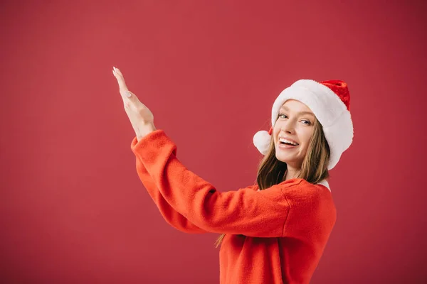 Attractive woman in santa hat and sweater pointing with hand isolated on red — Stock Photo