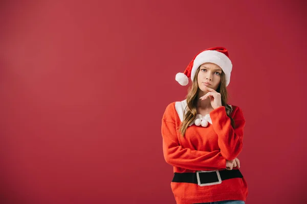 Attractive and pensive woman in santa hat and sweater looking at camera isolated on red — Stock Photo