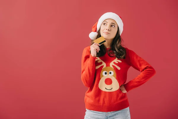 Attractive and pensive woman in santa hat and sweater holding credit card isolated on red — Stock Photo