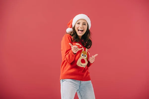 Attractive and smiling woman in santa hat and sweater pointing with fingers isolated on red — Stock Photo