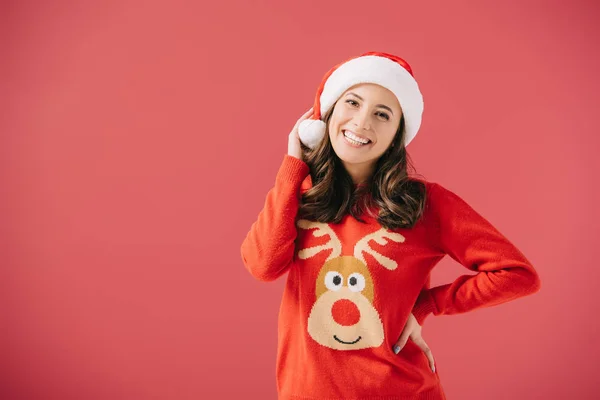 Atractiva y sonriente mujer en sombrero de santa y suéter mirando a la cámara aislada en rojo - foto de stock