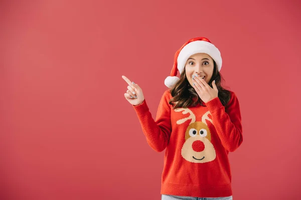 Femme attrayante et choquée dans le chapeau de Père Noël et pull pointant avec le doigt isolé sur rouge — Photo de stock