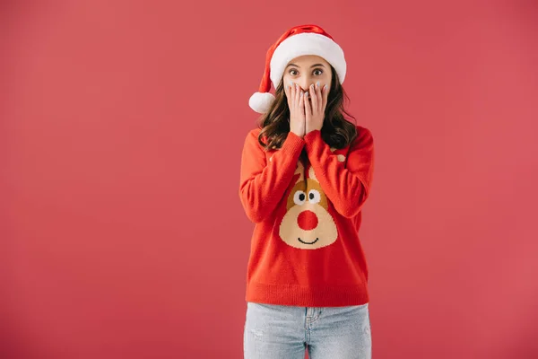 Attractive and shocked woman in santa hat and sweater looking at camera isolated on red — Stock Photo