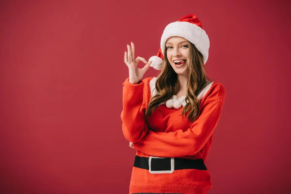 Attractive woman in santa hat and sweater showing ok gesture isolated on red — Stock Photo