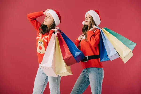 Attraktive Frauen in Pullovern und Weihnachtsmützen, die Einkaufstüten in der Hand halten und auf rotem Grund vereinzelt wegschauen — Stockfoto