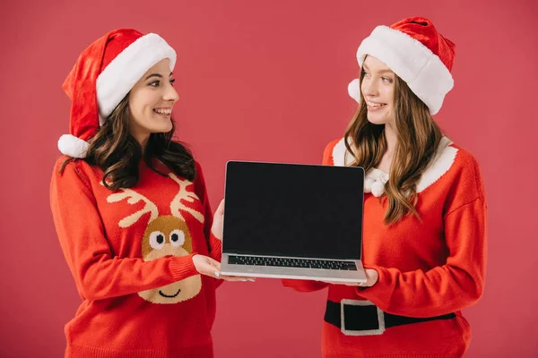 Mulheres atraentes e sorridentes em camisolas e laptop Santa segurando isolado no vermelho — Fotografia de Stock