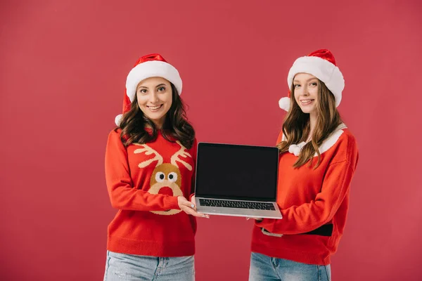 Привлекательные и улыбающиеся женщины в свитерах и Санта держа ноутбук изолирован на красный — стоковое фото