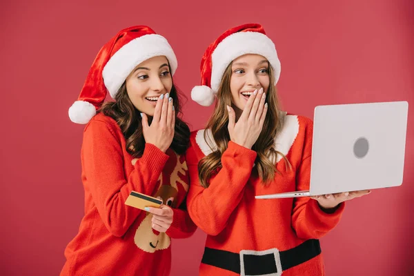 Attrayant et souriant femmes en chandails et Père Noël tenant ordinateur portable et carte de crédit isolé sur rouge — Photo de stock