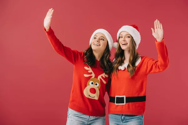 Donne attraenti e sorridenti in maglioni e cappelli di Babbo Natale che ondeggiano isolati sul rosso — Foto stock