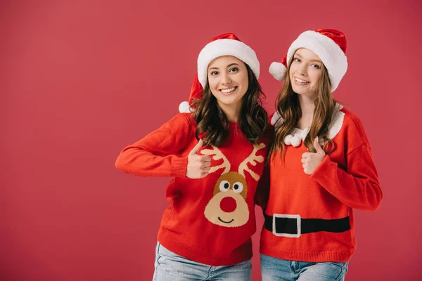 Attraente e sorridente donne in maglioni e cappelli di Babbo Natale mostrando pollici in su isolato sul rosso — Foto stock