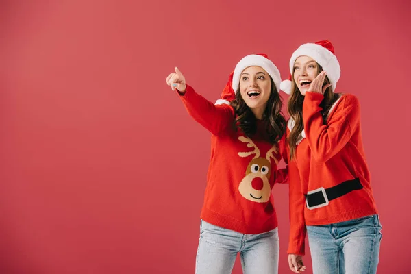 Attractive and smiling women in sweaters and santa hats pointing with finger isolated on red — Stock Photo