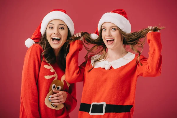 Attractive and smiling women in sweaters and santa hats playing with hair isolated on red — Stock Photo