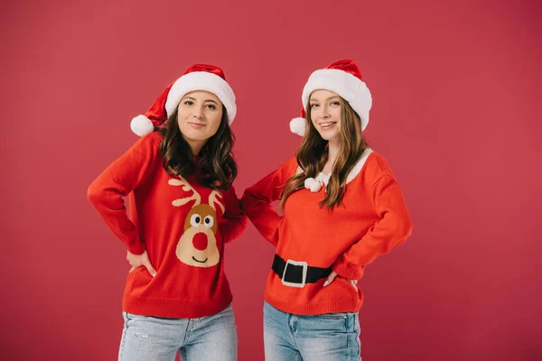 Donne attraenti e sorridenti in maglioni e cappelli di Babbo Natale con le mani sui fianchi guardando la fotocamera isolata sul rosso — Foto stock
