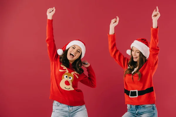 Attractive and smiling women in sweaters and santa hats with outstretched hands isolated on red — Stock Photo