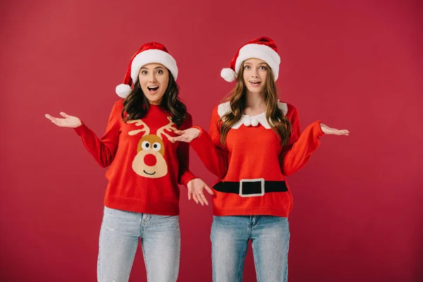 Femmes attirantes et choquées dans des pulls et des chapeaux de Père Noël avec les mains tendues isolées sur rouge — Photo de stock