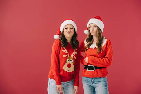 Attractive and smiling women in sweaters and santa hats making faces isolated on red — Stock Photo