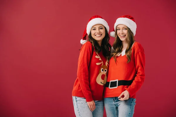 Mujeres atractivas y sonrientes en suéteres y sombreros de santa mirando a la cámara aislada en rojo - foto de stock