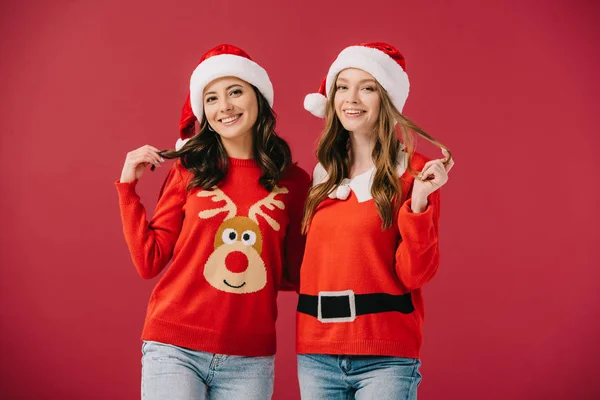 Donne attraenti e sorridenti in maglioni e cappelli di Babbo Natale guardando la fotocamera isolata sul rosso — Foto stock