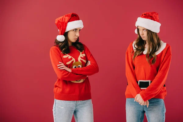 Attraente e offeso le donne in maglioni e cappelli di Babbo Natale guardando l'un l'altro isolato su rosso — Foto stock