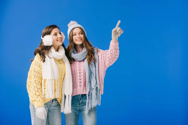 Attractive and smiling women in sweaters and scarves pointing with finger and looking away isolated on blue — Stock Photo
