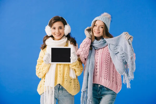 Mujeres atractivas y sonrientes en suéteres y bufandas que sostienen la tableta digital y mirando a la cámara aislada en azul - foto de stock