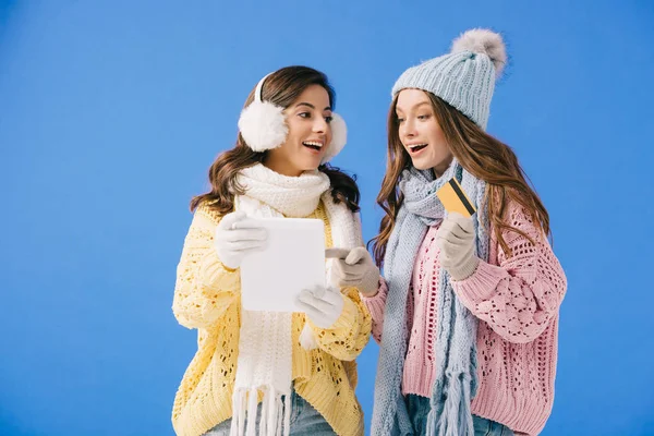 Mujeres atractivas y sonrientes en suéteres y bufandas que sostienen la tableta digital y mirando a la cámara aislada en azul - foto de stock