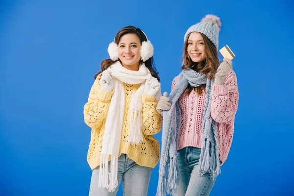 Attractive and smiling women in sweaters and scarves holding credit card isolated on blue — Stock Photo