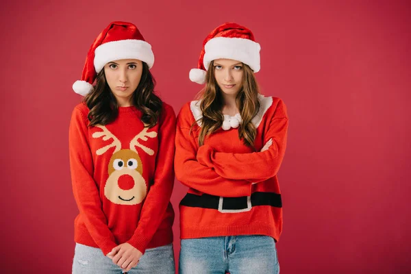 Mulheres atraentes e ofendidas em suéteres e chapéus de Papai Noel olhando para a câmera isolada no vermelho — Fotografia de Stock