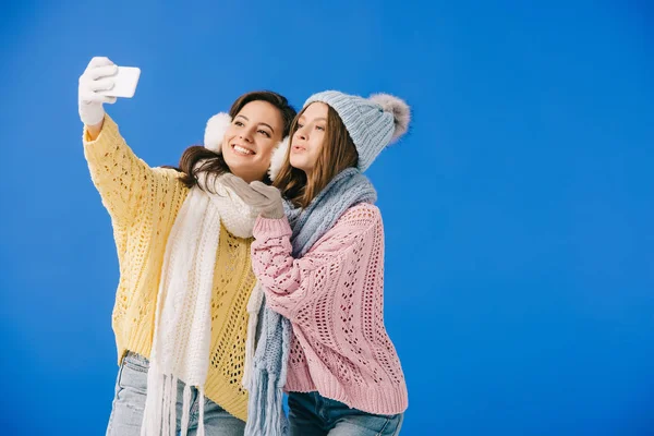 Mulheres atraentes e sorridentes em suéteres e cachecóis tomando selfie e soprando beijo isolado em azul — Fotografia de Stock