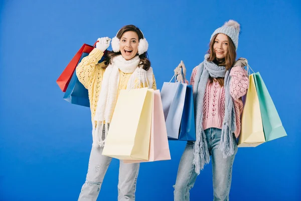 Attractive and smiling women in sweaters and scarves holding shopping bags isolated on blue — Stock Photo