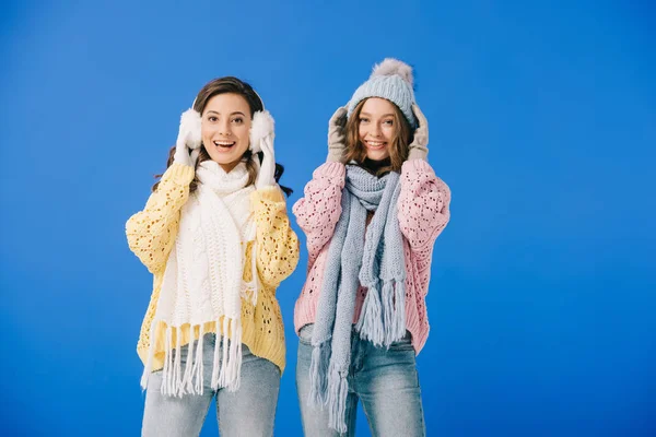 Mujeres atractivas y sonrientes en suéteres y bufandas mirando a la cámara aislada en azul - foto de stock