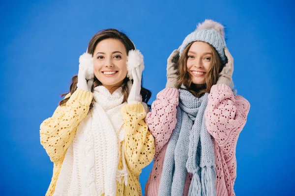 Mujeres atractivas y sonrientes en suéteres y bufandas mirando a la cámara aislada en azul - foto de stock