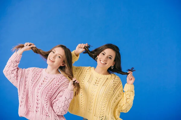 Привабливі і усміхнені жінки в светрах, які грають з волоссям, дивлячись на камеру ізольовані на синьому — стокове фото